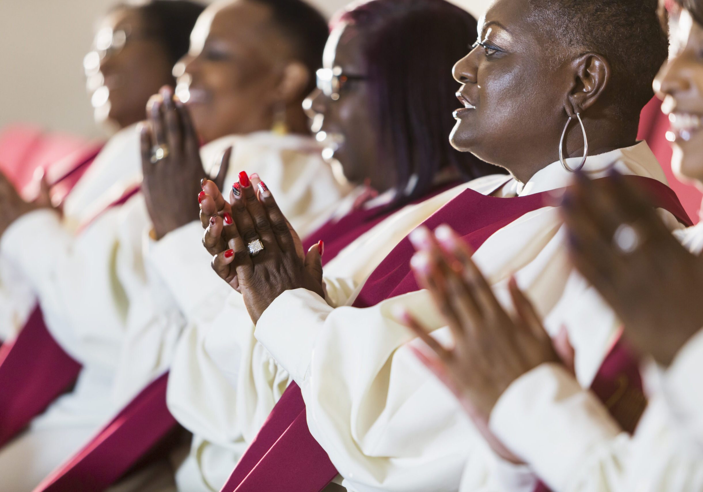 A group of mature black women in church robes, sitting in a row, clapping. They are members of the church choir listening to a sermon.